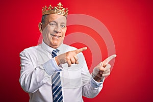 Senior handsome businessman wearing king crown standing over isolated red background smiling and looking at the camera pointing
