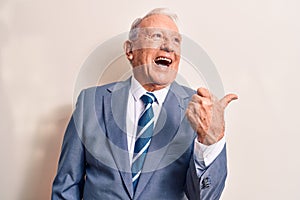 Senior handsome grey-haired businessman wearing elegant suit over white background pointing thumb up to the side smiling happy