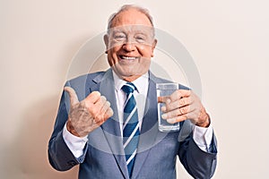 Senior handsome grey-haired businessman wearing suit drinking glass of water to refreshment pointing thumb up to the side smiling