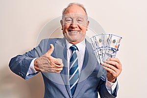 Senior handsome grey-haired businessman wearing suit holding bunch of dollars banknotes smiling happy and positive, thumb up doing