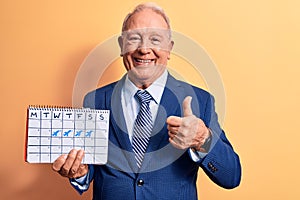 Senior handsome grey-haired businessman wearing suit holding business travel calendar smiling happy and positive, thumb up doing