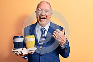 Senior handsome grey-haired businessman wearing suit holding cups of takeaway coffee pointing thumb up to the side smiling happy