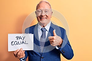 Senior handsome grey-haired businessman wearing suit holding paper with pay equal message smiling happy and positive, thumb up