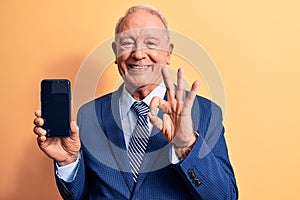 Senior handsome grey-haired businessman wearing suit holding smartphone showing screen doing ok sign with fingers, smiling