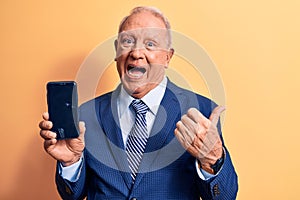 Senior handsome grey-haired businessman wearing suit holding smartphone showing screen pointing thumb up to the side smiling happy