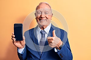 Senior handsome grey-haired businessman wearing suit holding smartphone showing screen smiling happy and positive, thumb up doing