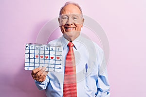 Senior handsome grey-haired businessman wearing tie holding cardio heart calendar looking positive and happy standing and smiling