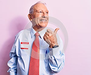 Senior handsome grey-haired businessman wearing tie and shirt with name presentation sticker pointing thumb up to the side smiling