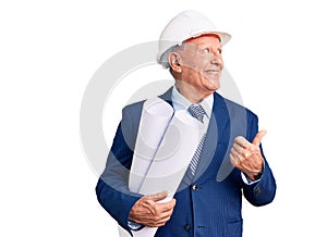 Senior handsome grey-haired man wearing architect hardhat holding blueprints pointing thumb up to the side smiling happy with open