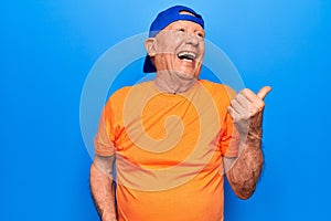 Senior handsome grey-haired man wearing casual t-shirt and cap over blue background pointing thumb up to the side smiling happy