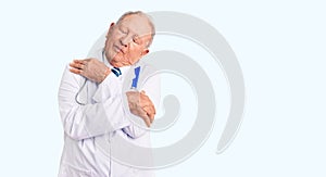 Senior handsome grey-haired man wearing doctor coat and stethoscope hugging oneself happy and positive, smiling confident