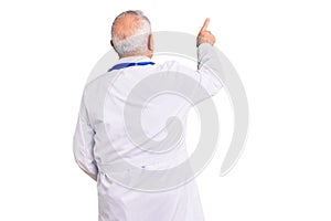 Senior handsome grey-haired man wearing doctor coat and stethoscope posing backwards pointing ahead with finger hand