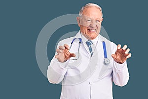 Senior handsome grey-haired man wearing doctor coat and stethoscope smiling funny doing claw gesture as cat, aggressive and sexy
