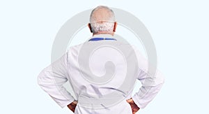 Senior handsome grey-haired man wearing doctor coat and stethoscope standing backwards looking away with arms on body