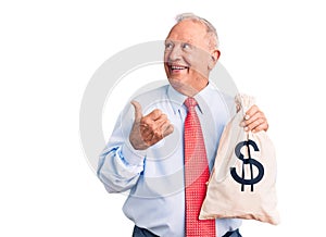 Senior handsome grey-haired man wearing elegant clothes holding money bag pointing thumb up to the side smiling happy with open
