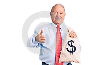 Senior handsome grey-haired man wearing elegant clothes holding money bag smiling happy and positive, thumb up doing excellent and