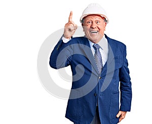 Senior handsome grey-haired man wearing suit and architect hardhat pointing finger up with successful idea