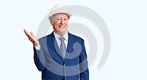 Senior handsome grey-haired man wearing suit and architect hardhat smiling cheerful presenting and pointing with palm of hand