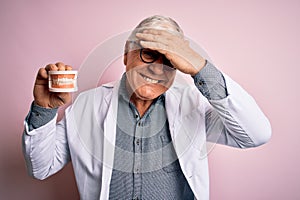 Senior handsome hoary dentist man wearing coat holding denture teeth over pink background stressed with hand on head, shocked with