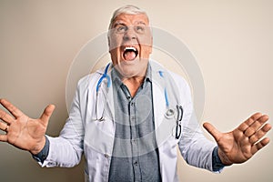 Senior handsome hoary doctor man wearing coat and stethoscope over white background crazy and mad shouting and yelling with