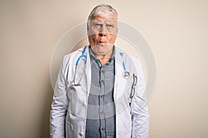 Senior handsome hoary doctor man wearing coat and stethoscope over white background making fish face with lips, crazy and comical