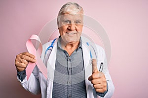 Senior handsome hoary doctor man wearing stethoscope holding pink cancer ribbon happy with big smile doing ok sign, thumb up with