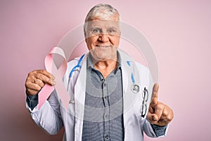 Senior handsome hoary doctor man wearing stethoscope holding pink cancer ribbon surprised with an idea or question pointing finger