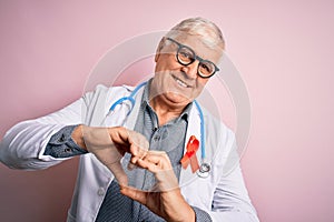 Senior handsome hoary doctor man wearing stethoscope and red HIV ribbon on coat smiling in love showing heart symbol and shape