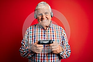 Senior handsome hoary gamer man playing video game using joystick and headphones with a happy face standing and smiling with a