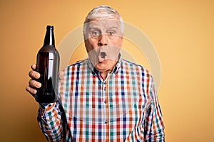 Senior handsome hoary man drinking bottle of beer standing over isolated yellow background scared in shock with a surprise face,