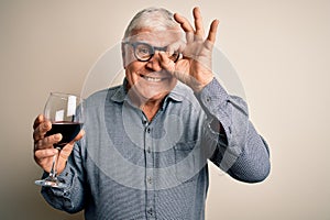 Senior handsome hoary man drinking glass of red wine over isolated white background with happy face smiling doing ok sign with