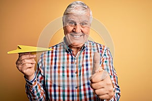 Senior handsome hoary man holding paper airplane standing over isolated yellow background happy with big smile doing ok sign,