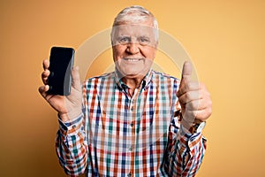 Senior handsome hoary man holding smartphone showing screen over yellow background happy with big smile doing ok sign, thumb up