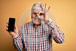 Senior handsome hoary man holding smartphone showing screen over yellow background with happy face smiling doing ok sign with hand