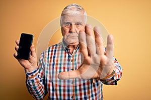 Senior handsome hoary man holding smartphone showing screen over yellow background with open hand doing stop sign with serious and