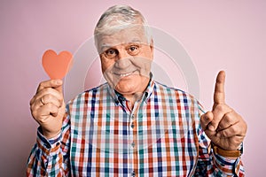 Senior handsome hoary romantic man holding red paper heart shape over pink background surprised with an idea or question pointing