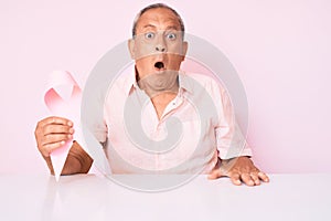 Senior handsome man with gray hair holding pink cancer ribbon sitting on the table scared and amazed with open mouth for surprise,