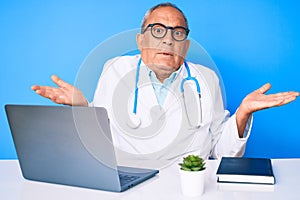 Senior handsome man with gray hair wearing doctor uniform working using computer laptop clueless and confused expression with arms