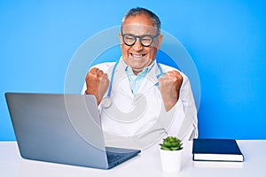Senior handsome man with gray hair wearing doctor uniform working using computer laptop excited for success with arms raised and