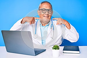 Senior handsome man with gray hair wearing doctor uniform working using computer laptop smiling cheerful showing and pointing with