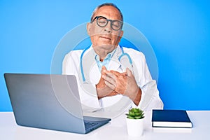 Senior handsome man with gray hair wearing doctor uniform working using computer laptop smiling with hands on chest with closed