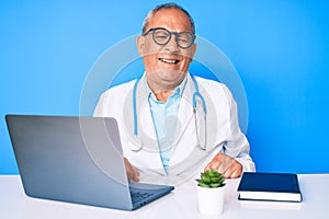 Senior handsome man with gray hair wearing doctor uniform working using computer laptop winking looking at the camera with sexy