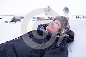 Senior handsome man in warm clothes and hat lying on snow and dreaming