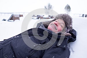 Senior handsome man in warm clothes and hat lying on snow and dreaming