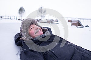 Senior handsome man in warm clothes and hat lying on snow and dreaming