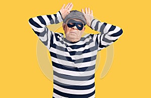 Senior handsome man wearing burglar mask and t-shirt doing bunny ears gesture with hands palms looking cynical and skeptical