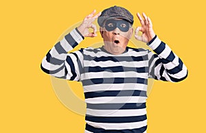 Senior handsome man wearing burglar mask and t-shirt looking surprised and shocked doing ok approval symbol with fingers