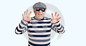 Senior handsome man wearing burglar mask and t-shirt smiling funny doing claw gesture as cat, aggressive and sexy expression