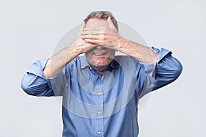 Senior happy man in blue shirt covering eyes with hands