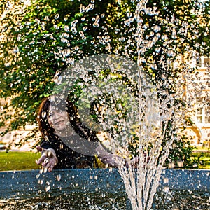 Senior happy woman by the fountain catches water droplets on a sparkling sunny summer day